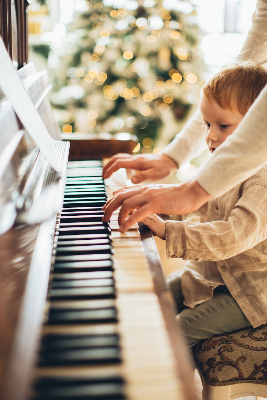 A young boy is playing Christmas music at the piano with a parent or teacher reaching around to help him
